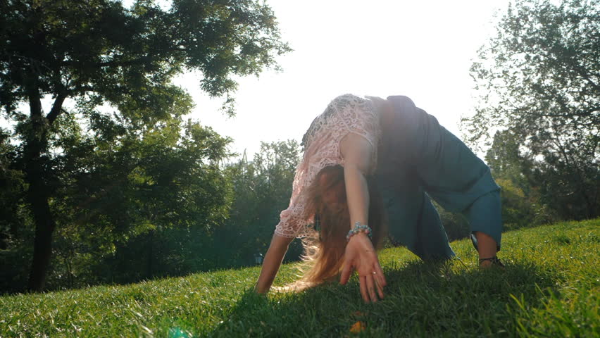 Portrait of young white woman in blue pants runner stretching legs in the park. Green grass, park, summer morning. Relaxing and meditating, Health concept. Slow motion.