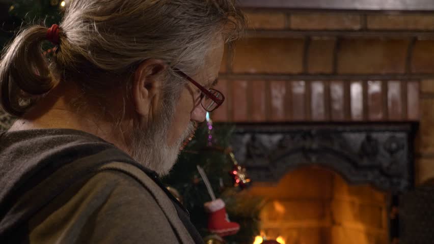 Senior man reading book near the fireplace and decorated Christmas tree at xmas eve, slow motion.