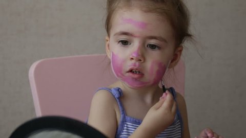 blonde three years old baby painting color stripes on her face looking at mirror