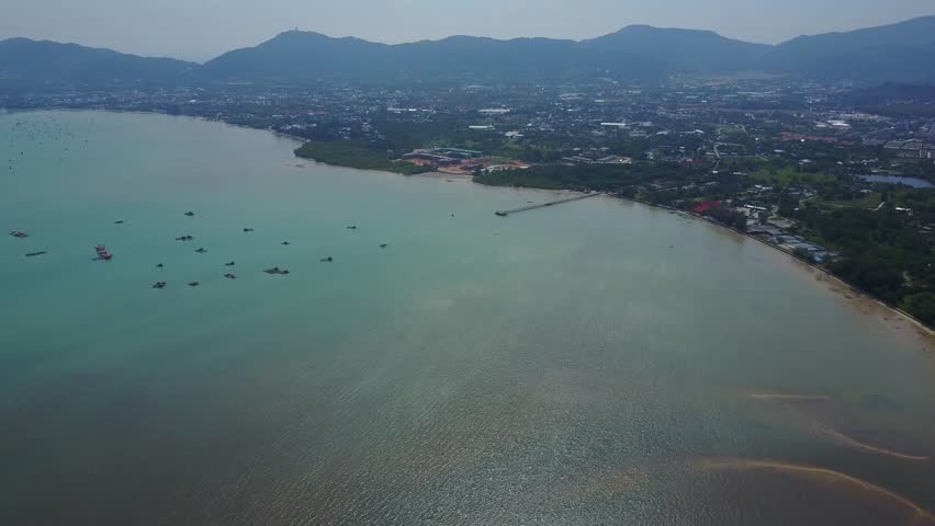 Chalong bay green and dark grey water, confluence of seawater with river flow, large town area on shore, small pearl farm floating constructions on left. Dim mountains silhouettes on horizon