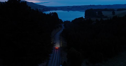 Aerial view car driving on country road at dusk through forest with headlights