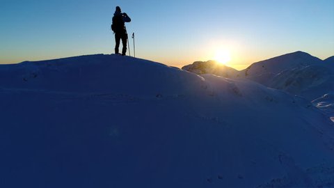 Aerial - Male mountaineer standing on top of snowy mountain and taking photos of beautiful winter scenery with his phone at sunset