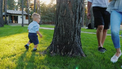 Happy parents play hide and seek with a baby boy. He runs happily behind them around the tree. Happy time with parents