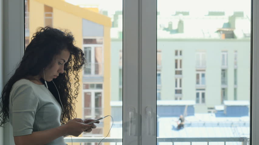 Curly brunette messaging with someone listening a music near the window