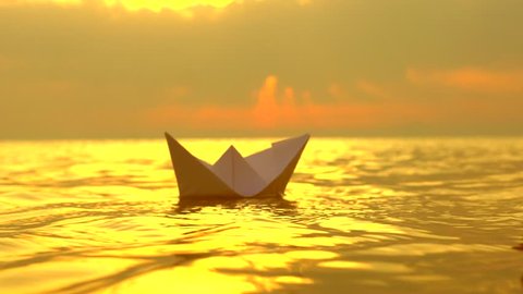 Kid with a paper boat in water over beautiful sunset. Little boy's hand puts paper ship on sea surface. Origami ship Sailing. Dreams, future, childhood, freedom or hope concept. Slow motion 4K
