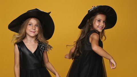 Little playful girls witches in decorated dresses and hats dancing and smiling isolated over orange