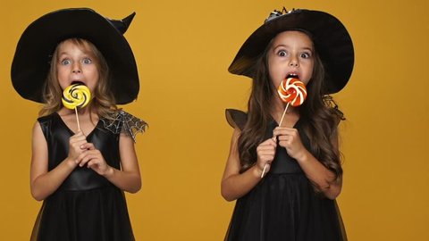 Little pretty girls witches in decorated dresses and hats eating lollipops isolated over orange