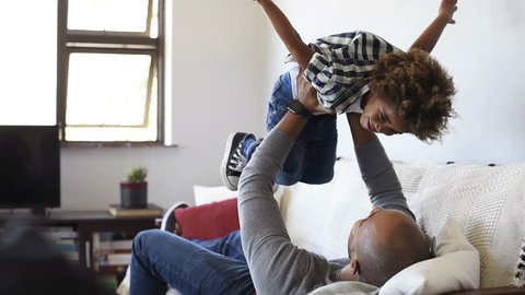 Cheerful young boy having fun with his father on sofa. Little african son playing with father on couch with open outstretched arms. Black child enjoying playing with dad at home.