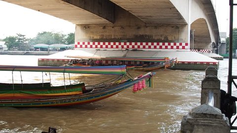 BANGKOK, THAILAND - SEPTEMBER 21, 2017: Wooden long-tailed boats with roof stopping, parking under Phra Pinklao concrete large bridge, on waving muddy brown water river, beside pier, bright white sky