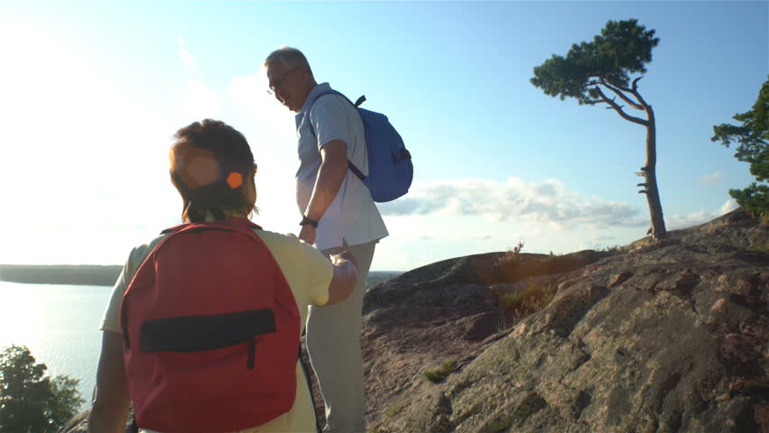 Elderly active couple climbing on the rock and admiring the sunrise, the sunset. Scandinavian landscape with sea and pine trees.