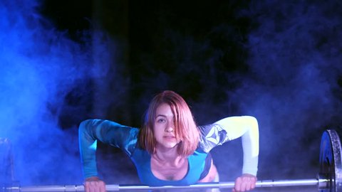 athletic girl is doing push ups with a barbell, At night, in light smoke, fog, in light of multicolored searchlights, in an old abandoned hangar, building