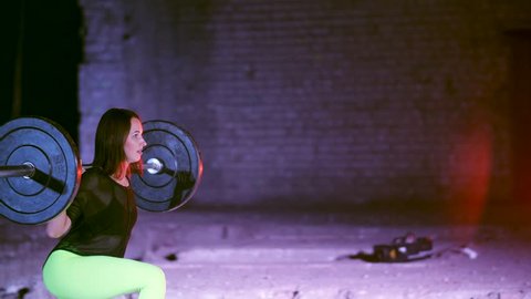 athletic young woman, raises the barbell, does sit-ups with the barbell, it's hard for her. At night, in light of multicolored searchlights, in light smoke, fog, in old abandoned building