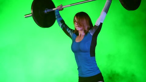 athletic young woman, raises the barbell, does sit-ups with the barbell, it's hard for her. At night, in light of multicolored searchlights, in light smoke, fog, in old abandoned building