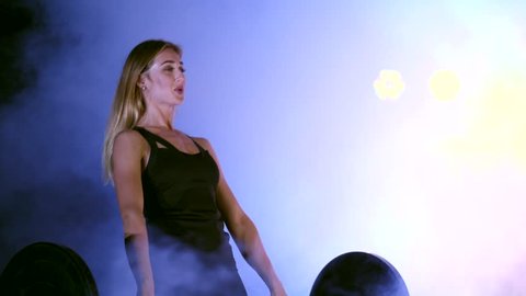 athletic, beautiful, sexy woman does exercise with the barbell, raises it. At night, in the light of multicolored searchlights, in light smoke, fog, in an old abandoned building.
