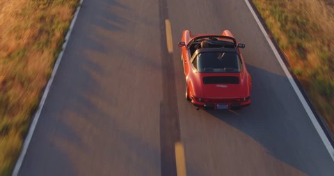 Aerial View Of Happy Couple Driving Vintage Convertible Sports Car Down Country Road At Sunset