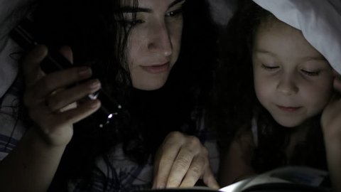 Mom and her little daughter are reading fairy tales for the night, they are wrapped in a warm blanket and immersed in the story of the plot. 4K.