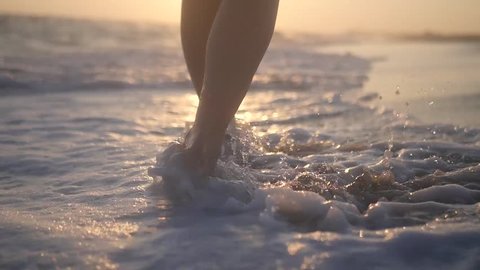Beautiful female feet walking along the seashore with waves and splashing against the backdrop of a beautiful sunset. HD, 1920x1080. slow motion.