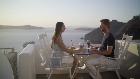 4k travel video honeymoon couple enjoying private dinner at sunset outside on terrace in santorini island overlooking the caldera cheering with red wine
