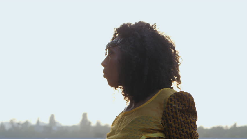 Portrait of young afro-american gypsy woman in colorful yellow traditional dress and silver crown on forehead smiling and posing close to camera. Sexy fashion girl with curly hair in slow motion.