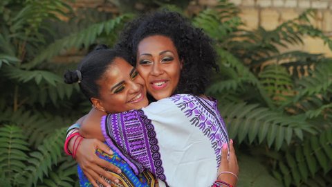 Two happy beautiful afro-american women friends hugging outdoor in park. Multi ethnic girls wearing colorful clothing enjoys the meeting and laughing in slow motion. Dolly shot.
