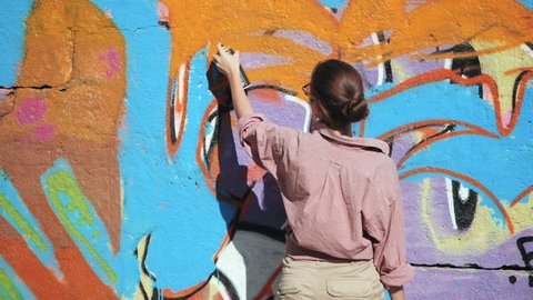 Beautiful Young Girl making a colorful graffiti with aerosol spray on urban street wall. Cinematic toned slow motion footage. Creative art. Talented student in glasses drawing picture.