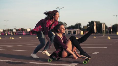 Smiling woman sitting on a longboard while her friend is pushing her behind and running during sunset. Enjoying life. Lens flare. Slowmotion shot