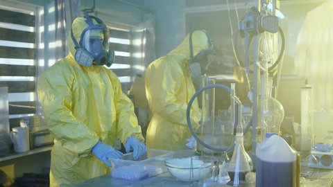 In the Underground Laboratory Two Clandestine Chemists Wearing Protective Masks and Coveralls Pack Bags Full of Crystal Meth into Boxes. Equipment. They Squat in an Abandoned Building. 4K UHD.