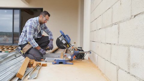 handsome young man carpenter using a circular saw while installing wood floor terrace outdoor in new house construction site