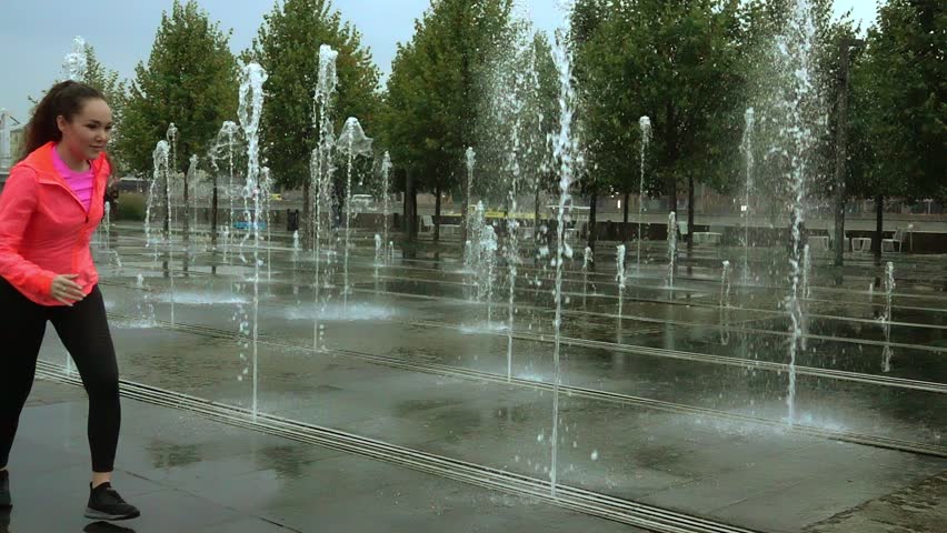 Young mixed race athletic woman running in the rain near city park fountains, super slow motion video