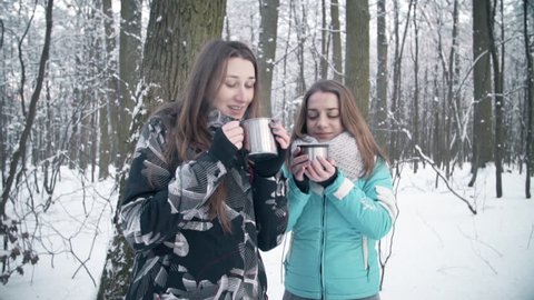 Girls drinking hot tea outdoors in winter woods. Girls together are drinking cup of hot tea and smiling. Girls drinking hot tea or coffee outdoors in winter woods.