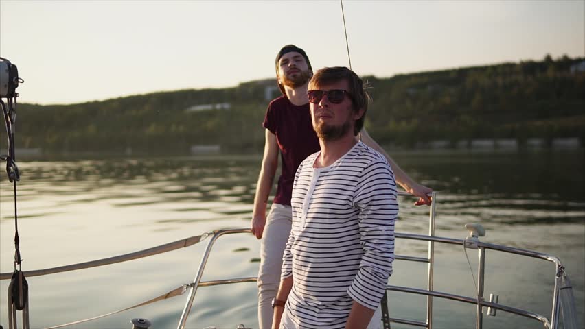 Young friends rest on the yacht in the evening, friends are engaged in sailing in the summer, men look at the sunset and enjoy the sea smoothness behind the boat of their sea vessel