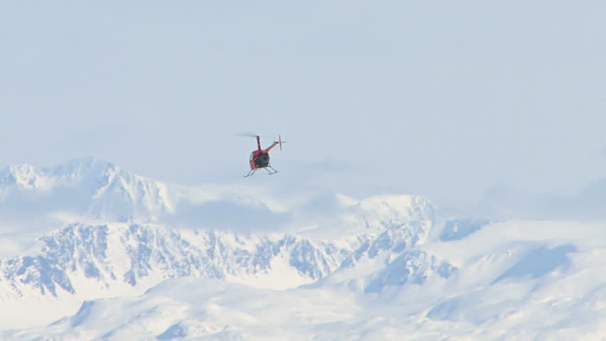 Red 2-man helicopter (Robinson R22) in the distance, flying away toward snowy