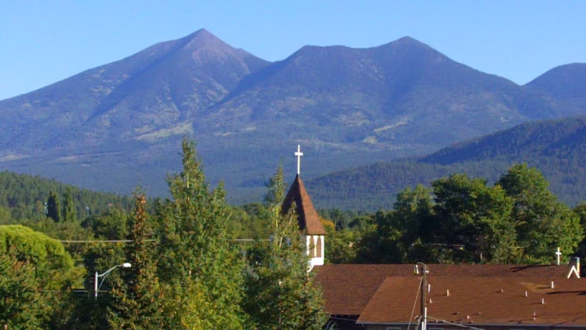 A church steeple and pine trees reach upward against a backdrop of mountains in