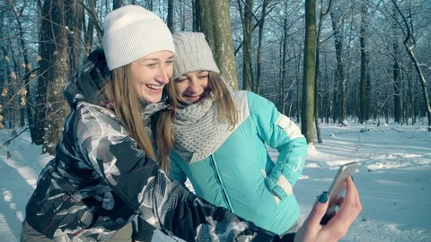 Two girls taking selfie in winter forest. Girls happily take selfie in winter forest, laughing and smiling. Girl Hold Smart Phone Camera Taking Selfie Photo Two Young Woman Snow Forest Outdoor Winter 