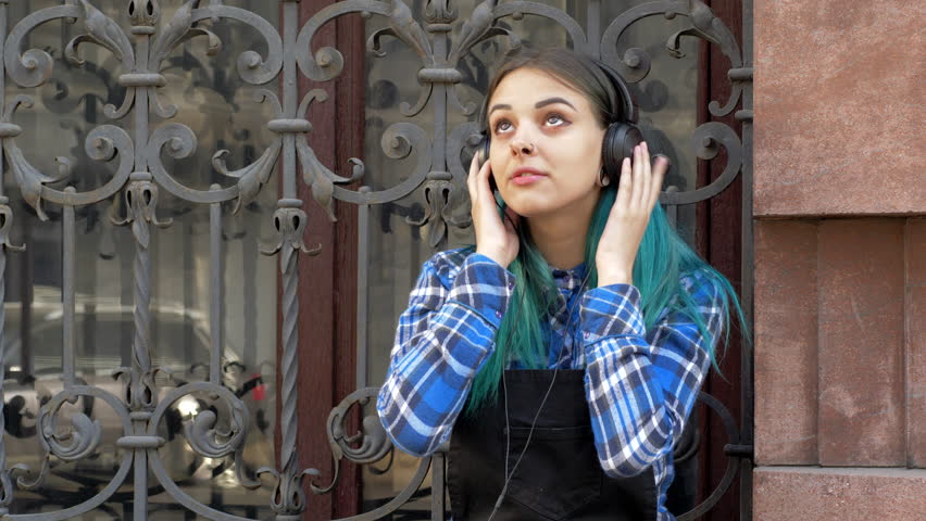 Street hipster girl listening music with brown headphones.Teen girl with blue dyed hair,piercing in nose,violet lenses,unusual hairstyle sitting near historic building. 4k