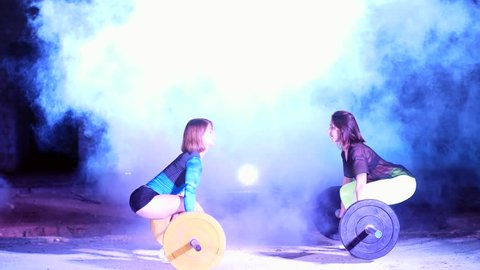 Two athletic girls, athletes, doing exercises with the barbell. At night, in the light of searchlights, a stobascope, in light smoke, fog, in an old abandoned building.
