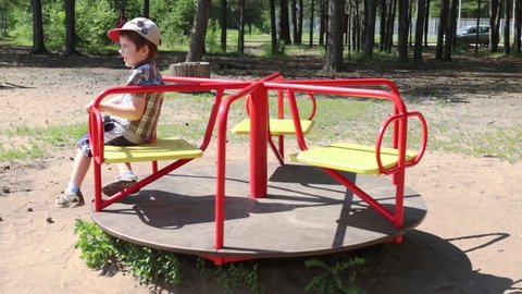 Happy little boy waves hand on carousel in children playground in park
