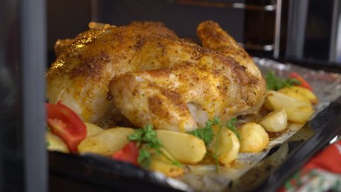 Cooking whole roasted chicken with vegetables baked in the oven. Taking out crispy chicken on a baking sheet from the oven. Roast chicken with potatoes tomato slices herbs cooked in electric cooker