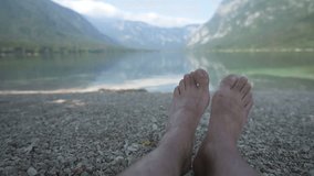 Barefoot caucasian male relaxing by lake water, selective focus on man's feet lying on lakeshore. Summer vacation and travel destination scenic. Flat profile ungraded footage with selective focus