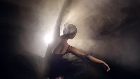 ballerina with hair in a bun is dancing on a scene, against bright light of soffit and smokescreen