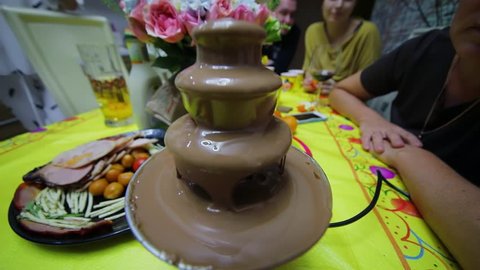 People sit around table with chocolate waterfall and food during birthday party