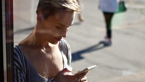 A girl is looking into her cell phone. The video is filmed through a cafe showcase. A woman communicates in social networks on a sunny day.