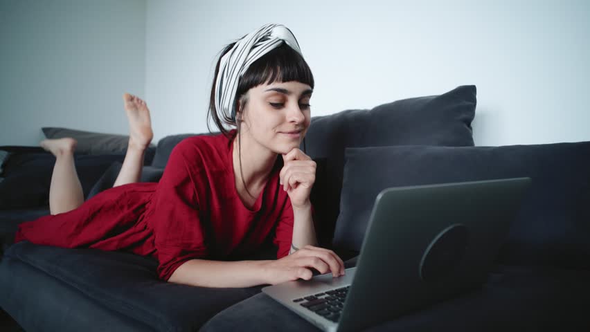 Cheerful, happy optimistic young millennial hipster woman or housewife relaxed on sofa lays in comfortable clothes, purchases things online through web stores, uses credit card