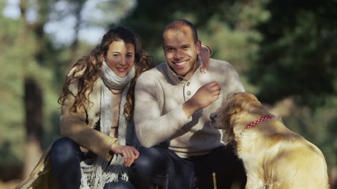 Portrait of an attractive couple of mixed ethnicity and their loveable dog. They are sitting in a forest clearing with dappled sunlight shining on their faces. 