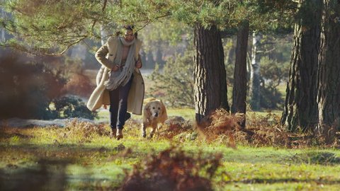 Attractive young couple are exercising their dog and running through a sunlit forest on a cool bright autumn morning. In slow motion.