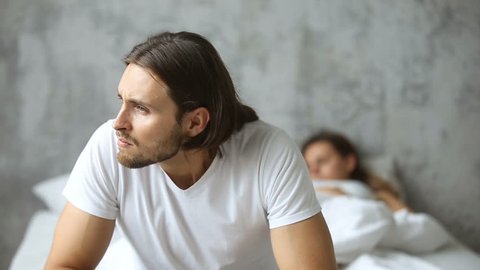 Thoughtful worried man sitting on the side of bed with sleeping woman at background, doubtful upset husband thinking of breaking up divorce, feeling unsure frustrated obsessed about family problem