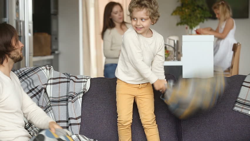 Little cute boy son having funny pillow fight with dad on couch in living room while mom and girl daughter cooking at background, happy couple family spending time with small kids at home concept Royalty-Free Stock Footage #31060639