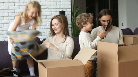 Happy excited family with two children unpacking boxes moving into new home concept, small kids helping parents with belongings in cardboards putting cushion on sofa in living room after relocation 