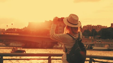 Woman tourist in Paris. Traveller female in hat, shooting on camera in her phone beautiful sunset near the Seine river.