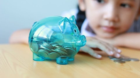 Asian little girl in Thai kindergarten student uniform putting money coin into clear piggy bank on wooden table metaphor money saving for education concept shallow depth of field select focus on pig